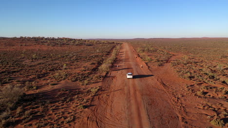 Aerial:-Drone-shot-closely-following-a-white-vehicle-as-it-drives-along-a-dusty-red-outback-road,-near-Broken-Hill,-Australia