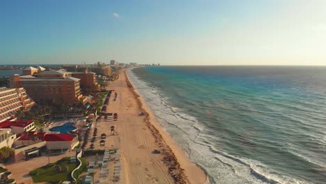 Tropical-sea-waves-crashing-on-white-sandy-shore-with-a-blue-sky,-golden-sun-rays-shine-on-turquoise-sea-next-to-Hotel-Zone-resorts---Laguna-Nichupté-in-Cancun,-Mexico---Drone-panning