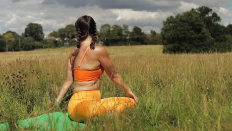 Woman-sitting-in-long-grass-field-doing-yoga,-stretching-arms-up-to-side-bend-to-easy-pose-bound-hands