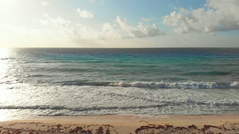Tropical-sea-waves-crashing-onto-the-white-sandy-shore-next-to-seaweed,-while-golden-sun-rays-shine-through-the-luminous-turquoise-sea-in-Cancun,-Mexico-with-white-clouds-in-a-blue-sky---4K-drone