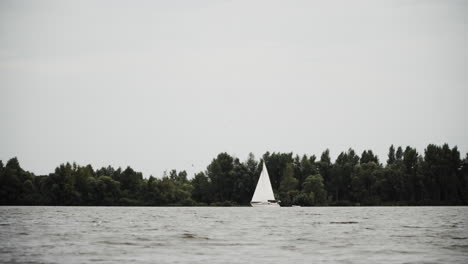 Ein-Segelboot-Segelt-Sanft-über-Den-Ruhigen-Fluss-In-Den-Niederlanden-Unter-Dem-Dramatischen-Himmel-Mit-üppigen-Bäumen-Im-Hintergrund---Weitschuss