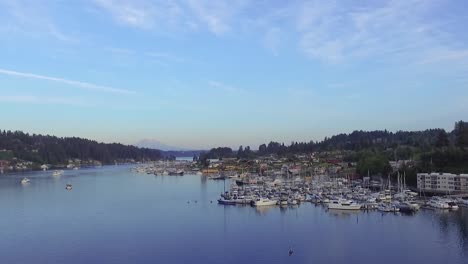 Sailboats-And-Yachts-Docked-At-The-Gig-Harbor-Marina-And-Boatyard-In-Pierce-County,-Washington-With-A-Kayaker-Paddling-Over-The-Calm-Puget-Sound-Bay---wide-aerial-shot