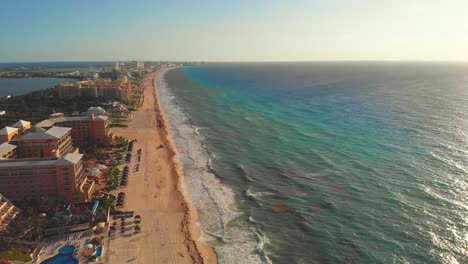 Drone-flying-towards-tropical-sea-waves-crashing-on-white-sandy-shore-with-a-blue-sky,-golden-sun-rays-shine-on-turquoise-sea---Hotel-Zone-resorts---Laguna-Nichupté-in-Cancun,-Mexico