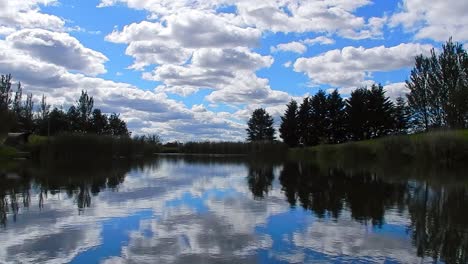 Vibrant-blue-lake-water-mirror-reflection-of-bright-clouds-and-trees-in-sky-landscape