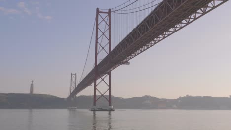 A-view-of-de-Abril-Bridge-where-a-boat-with-people-is-floating-on-the-Tagus-river,-Lisbon,-Portugal