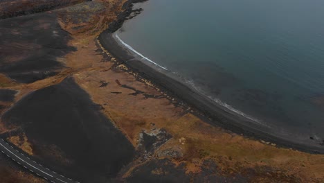 Epic-aerial-top-view-above-icelandic-road-by-black-sand-coastline,-waves-crashing-against-shore,-day