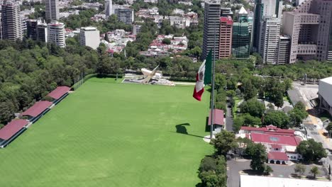 Flight-above-empty,-desolate,-deserted-and-closed-Campo-Marte-green-grass-venue-with-patriotic-Mexican-flag-waving-in-wind,-Mexico-city,-covid-19-pandemic,-lockdown-and-quarantine,-overhead-aerial