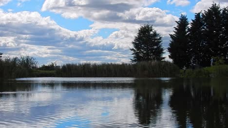 People-on-banks-of-blue-lake-water-mirror-reflection-bright-scenic-cloudy-sky