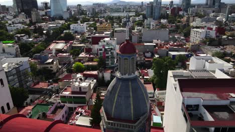 Mixcoac-church-tower,-steeple,-dome,-and-steel-cross-in-downtown-Mexico-city-center-buildings,-red-roofs-and-skyline,-rising-tilt-down-circle-aerial