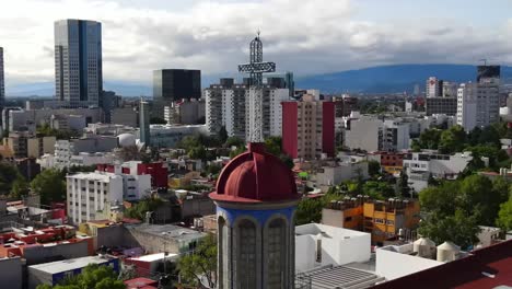 Vertigo-effect-of-church-tower,-steeple,-red-dome,-and-steel-cross-in-downtown-Mexico-city-center-buildings,-skyscrapers-and-skyline-with-mountain-range-in-background-on-cloudy-day,-aerial-zoom-in