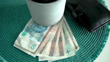 Schwenken-Sie-über-Zerknitterte-Polnische-Zloty-Noten,-Die-Unter-Einem-Weißen-Blumentopf-Neben-Einer-Schwarzen-Lederbrieftasche-Auf-Einem-Grünen-Tischset-Platziert-Sind