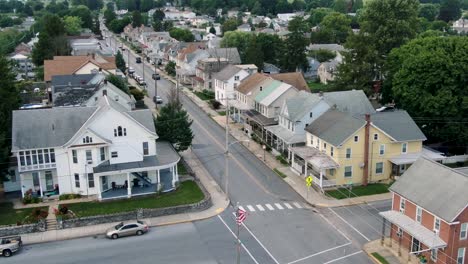 Establishing-shot,-aerial-of-Anytown-USA,-small-town-in-America-with-American-flag-flying,-United-States-community-neighborhood-homes,-historic-colonial-houses-line-street-as-traffic-passes-by,-summer