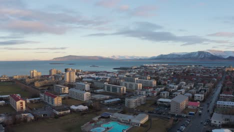 Rotating-aerial-view-of-urbanized-and-modern-city-Reykjavik,-capital-of-Iceland,-surrounded-by-snow-capped-mountains-under-a-cloudy-sky