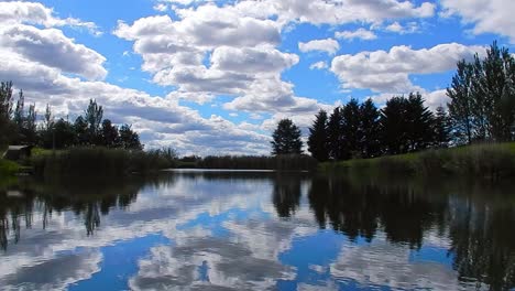 Vibrant-blue-lake-water-mirror-reflection-of-bright-clouds-and-trees-in-blue-sky