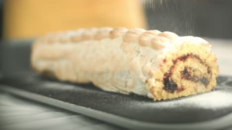 Close-up-of-a-swiss-roll-with-flambeed-egg-white-cream-while-powdered-sugar-is-put-on-top-of-the-roll-laying-on-a-plate