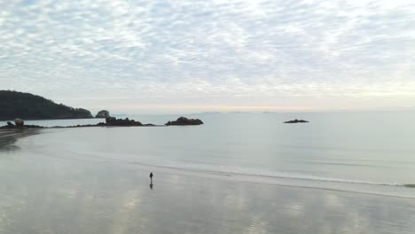 Person-walking-alone-on-empty-coastal-bay-beach-at-sunset,-aerial-arc-shot