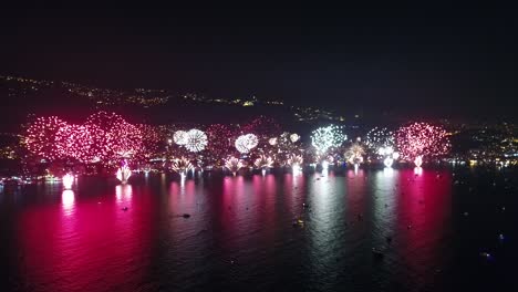Fireworks-lighting-up-city-by-the-sea-coast,-colorful-light-reflection-on-sea-water,-new-year-celebration,-static-aerial-drone-view,-night