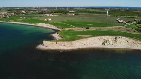 Aerial-of-Danish-coastline-in-the-summer-with-a-small-cliff-and-green-fields-with-giant-windmills-and-a-beautiful-blue-sky