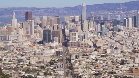 Top-aerial-view-of-the-skyline-of-Downtown-San-Francisco,-California---a-man-hikes-down-the-Twin-Peaks-mountain
