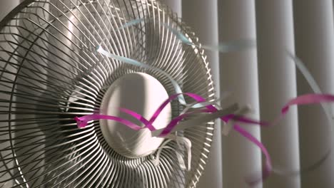 Closeup,-static-shot-of-a-rotating-fan-with-colorful-ribbons-on-the-grill,-in-the-background-closed-window-blinds-letting-sunlight