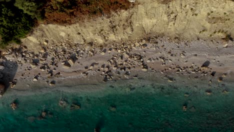 Rocky-beach-with-sand-and-cliffs-washed-by-emerald-clean-sea-water-at-golden-hour-in-Mediterranean,-top-down-aerial