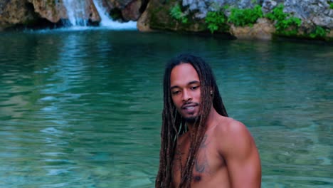 Afro-American-Man-With-Long-Braided-Hair-And-Tattoo-On-The-Left-Chest-Singing-In-The-River-With-Waterfall-In-The-Background---medium-close-up,-slow-motion