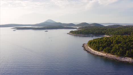 View-Of-The-Majestic-Mountain-In-The-Island-Of-Cres-From-The-Lush-Green-Paradise-Island-Of-Losinj-In-Croatia-With-A-Sailboat-Moored-Over-The-Adriatic-Sea---aerial-drone
