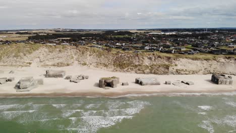 Aerial-of-beautiful-white-sand-beach-with-old-abandoned-German-bunkers-in-the-sand-wile-waves-crashes-and-houses-in-the-distance