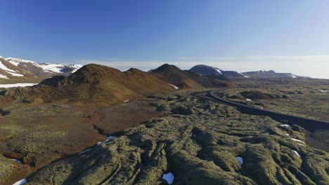 Volcanic-Landscape-With-Lava-Field-Covered-By-Moss-And-Snow-Mountain-With-Blue-Sky-At-The-Background-In-Iceland