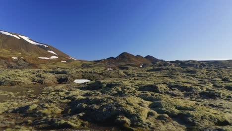 Lava-Field-Covered-By-Green-Moss-And-Snow-Mountain-With-Blue-Sky-At-The-Background-In-Iceland