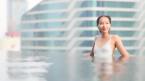 An-attractive-woman-relaxing-at-a-Hotel-with-the-city-skyline-of-Bangkok,-Thailand,-While-leaning-on-the-edge-of-a-rooftop-resort-infinity-pool
