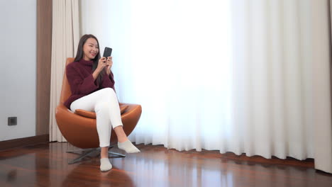 Ein-Junges-Asiatisches-Mädchen,-Das-Auf-Einem-Modern-Aussehenden-Sessel-Neben-Dem-Fenster-Sitzt-Und-Lächelnd-Auf-Ihrem-Mobiltelefon-Plaudert