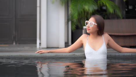 A-young-attractive-Asian-woman-in-a-one-piece-bathing-suit-leans-back-on-her-arms-on-the-edge-of-the-outdoor-swimming-pool,-slow-motion