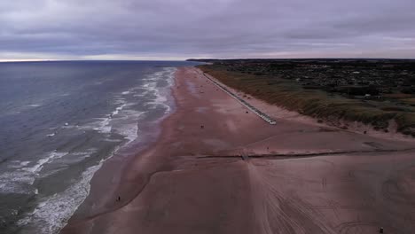Luftaufnahme-Der-Dänischen-Küste-Mit-Wunderschönem-Sandstrand,-Wellen-Und-Einer-Langen-Reihe-Kleiner-Strandhäuser-Bei-Einem-Wolkigen-Sonnenuntergang