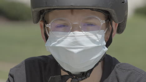 Portrait-of-teenage-boy-outside-wearing-glasses,-a-bike-helmet-and-a-medical-mask