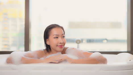 Close-up-on-an-Asian-woman-taking-bath-with-foam-looking-aside-and-smiling-at-the-hotel-resort