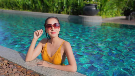 A-pretty-young-woman-fresh-from-a-dip-in-the-pool-leans-along-the-pool-edge-and-adjust-her-sunglasses