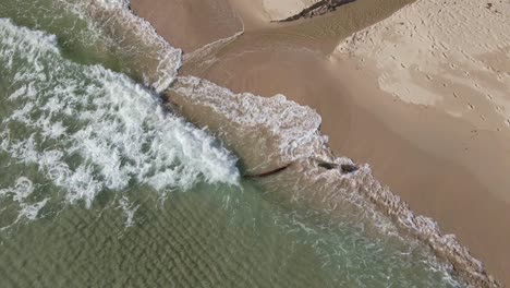 Aerial-drone-static-shot-of-clear-ocean-waves-crashing-slowly-on-a-white-sand-beach-with-driftwood