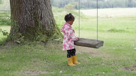 Lonely-adorable-little-African-girl-in-raincoat-waiting-to-play-near-swing,-static,-full-shot,-day
