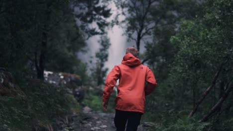 Slow-motion-shot-of-man-wearing-a-red-jacket-running-towards-a-big-waterfall-in-a-forest