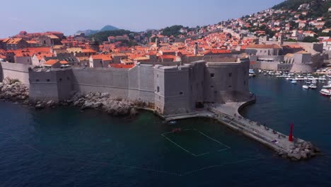 Aerial-reveal-flying-backwards-out-of-harbour-and-Old-Town-city-walls-of-Dubrovnik,-Croatia-with-mountains-and-sea-in-4k