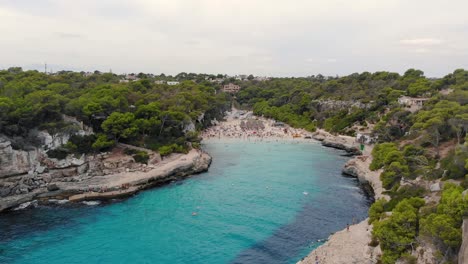 Aerial-Over-Turquoise-Waters-With-View-Of-Busy-Cala-d'Or-Beach