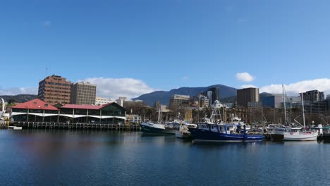 Timelapse-during-clear-sunny-day-at-small-old-dock-with-boats-and-yachts-tied-up-and-building-and-mountains-in-foreground-as-clouds-pass-by