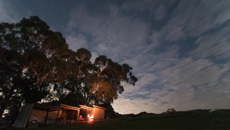 A-zoomed-in-time-lapse-of-a-man-having-a-camp-fire-by-a-shed-at-night-under-some-gum-trees-with-clouds-rolling-past-and-the-night-time-stars-in-the-background