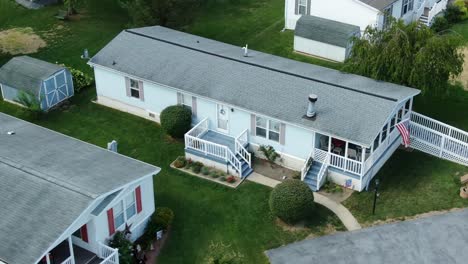 Typical-mobile-home-in-mobile-trailer-home-park,-American-flag,-blue-siding-and-gray-shutters,-white-deck-and-porch-railing-handicapped-accessible-disability-ramp-entrance-in-USA,-United-States-aerial