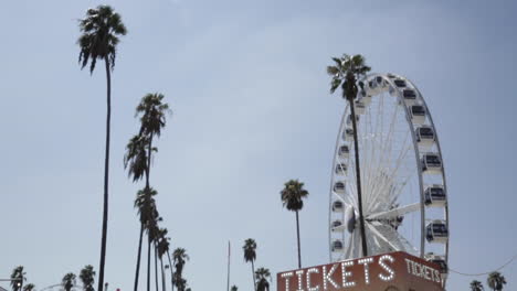 Empty-Ferris-Wheel-Spins,-Classic-Vintage-Tickets-Counter,-Palm-Trees,-Slow-Motion-Low-Angle