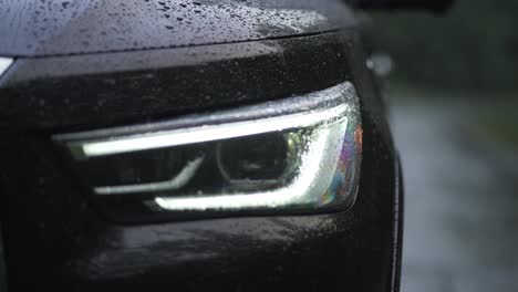 Schwarzer-High-End-Auto-LED-Scheinwerfer,-Der-Sich-Im-Regen-Einschaltet,-Wenn-Sich-Tautropfen-Auf-Der-Motorhaube-Bilden,-Extreme-Nahaufnahme