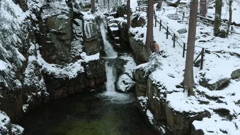 Aerial-view-of-a-person-jumping-in-a-icy-river,-at-a-waterfall,-in-middle-of-a-snowy-forest,-dark,-overcast,-winter-day---dolly,-drone-shot