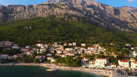 Aerial-view-flying-backwards-out-of-beach-revealing-huge-mountain-and-sea-during-sunset-in-Baska-Voda,-Croatia-in-4k