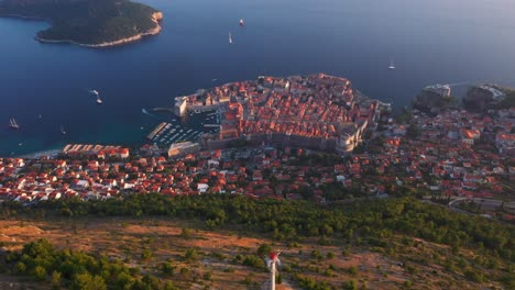Aerial-view-panning-down-from-sea-and-old-town-of-Dubrovnik,-Croatia-durning-sunset-revealing-mountains-and-cable-car-in-4k